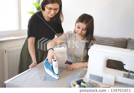 Young girl practices sewing skills at home with assistance. Girl and mom work together at a table with a sewing machine. Young girl practices sewing skills at home with assistance. Girl and mom work together at a table with a sewing machine. 139701851