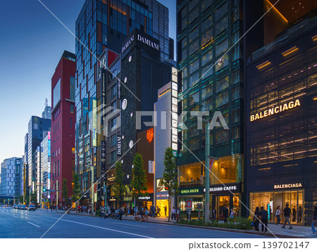 Tokyo, Japan - Oct 14 2024, panoramic view of main street of Ginza district, tall glass facades of skyscrapers along which people walk, cars drive along road, in evening with illumination, Tokyo Japan 139702147