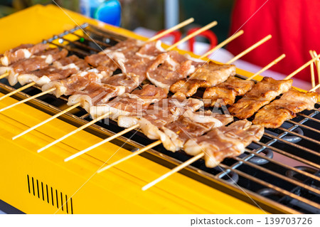 The shot of a cook grill Moo Ping or grilled pork skewers in Thai style on the yellow electric barbeque grill at the restaurant. The shot of a cook grill Moo Ping or grilled pork skewers in Thai style on the yellow electric barbeque grill at the restaurant. 139703726