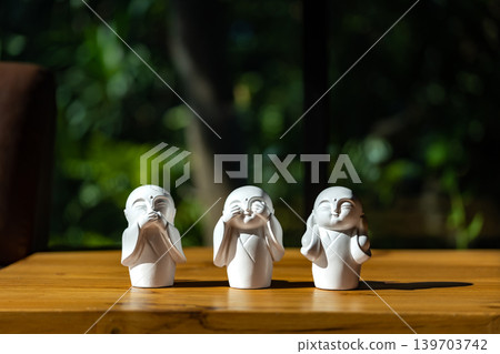 The white ceramic statue of three little monk See, Hear, Speak no evil set for home decoration placed on the wooden table over the black wall. The white ceramic statue of three little monk See, Hear, Speak no evil set for home decoration placed on the wooden table over the black wall. 139703742