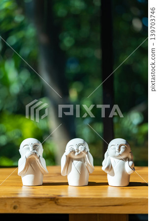 Three white monk figurines inspired by the Three Wise Monkeys philosophy are displayed as minimal zen decoration on a wooden tabletop with peaceful spiritual atmosphere. 139703746
