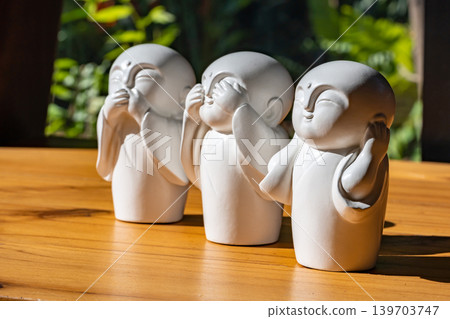 Three white monk figurines inspired by the Three Wise Monkeys philosophy are displayed as minimal zen decoration on a wooden tabletop with peaceful spiritual atmosphere. Three white monk figurines inspired by the Three Wise Monkeys philosophy are displayed as minimal zen decoration on a wooden tabletop with peaceful spiritual atmosphere. 139703747