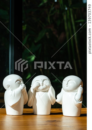 Three white monk figurines inspired by the Three Wise Monkeys philosophy are displayed as minimal zen decoration on a wooden tabletop with peaceful spiritual atmosphere. Three white monk figurines inspired by the Three Wise Monkeys philosophy are displayed as minimal zen decoration on a wooden tabletop with peaceful spiritual atmosphere. 139703749