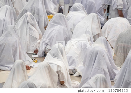 Hundreds of people sitting together covered with white fabric during a traditional spiritual ritual. Symbolic mass gathering expressing unity, faith, purity, and ceremonial devotion. Hundreds of people sitting together covered with white fabric during a traditional spiritual ritual. Symbolic mass gathering expressing unity, faith, purity, and ceremonial devotion. 139704323