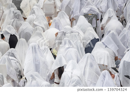 Hundreds of people sitting together covered with white fabric during a traditional spiritual ritual. Symbolic mass gathering expressing unity, faith, purity, and ceremonial devotion. Hundreds of people sitting together covered with white fabric during a traditional spiritual ritual. Symbolic mass gathering expressing unity, faith, purity, and ceremonial devotion. 139704324