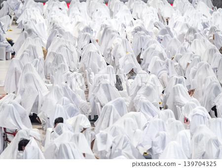 Hundreds of people sitting together covered with white fabric during a traditional spiritual ritual. Symbolic mass gathering expressing unity, faith, purity, and ceremonial devotion. Hundreds of people sitting together covered with white fabric during a traditional spiritual ritual. Symbolic mass gathering expressing unity, faith, purity, and ceremonial devotion. 139704325