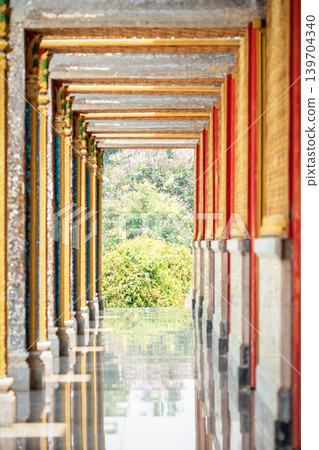 Long perspective corridor inside a Thai temple featuring golden frames, colorful columns, and symmetrical Buddhist architectural details. 139704340