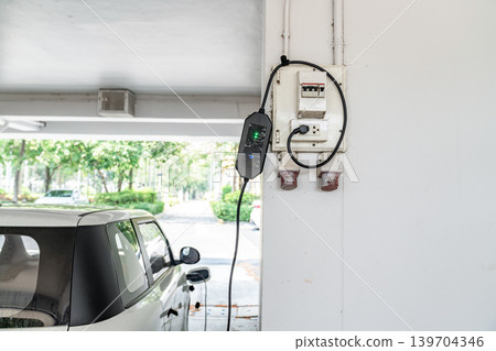 Portable electric vehicle charger connected to wall power outlet in parking area, representing sustainable transportation and clean energy technology. Portable electric vehicle charger connected to wall power outlet in parking area, representing sustainable transportation and clean energy technology. 139704346