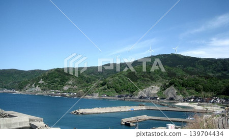 A landscape with a blue sky, a blue sea, lush green mountains, and white wind turbines. A landscape with a blue sky, a blue sea, lush green mountains, and white wind turbines. 139704434