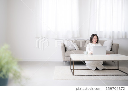 A woman using her laptop at home, studying for qualifications and considering investments; a wide-angle full-body shot of her lifestyle; copy space on the left. A woman using her laptop at home, studying for qualifications and considering investments; a wide-angle full-body shot of her lifestyle; copy space on the left. 139704872