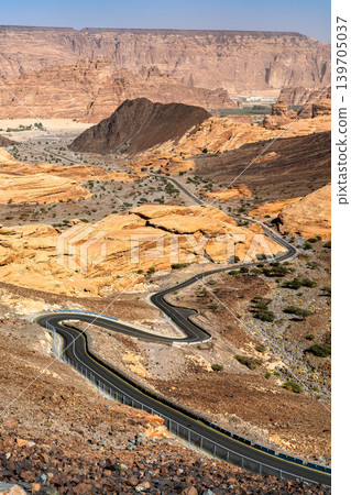 A winding asphalt road snakes through the dramatic sandstone rock formations and desert canyon landscape of AlUla, Saudi Arabia. 139705037