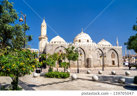 The historic stone facade and white domes of the Mosque of Al Ghamama stand against a clear blue sky in the holy city of Madinah, Saudi Arabia. 139705047