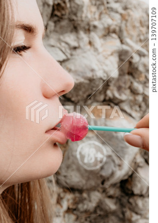 Woman lollipop candy. Close up profile of a young woman enjoying a pink lollipop against a textured rock wall background. Woman lollipop candy. Close up profile of a young woman enjoying a pink lollipop against a textured rock wall background. 139707109