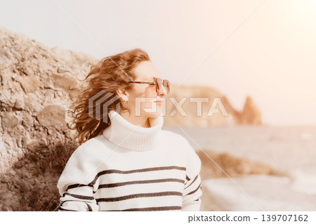 Woman beach vacation standing by rocks in a striped sweater and sunglasses enjoying the coastal scenery 139707162
