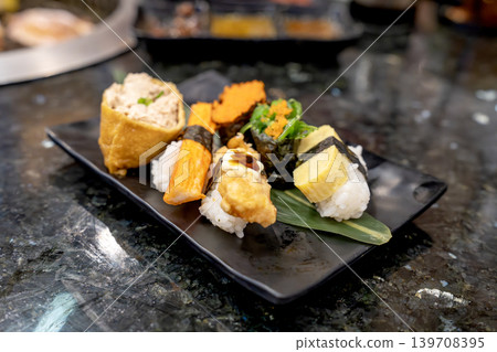 Assorted Japanese sushi platter featuring tamago, crab stick, inari tofu, and seaweed salad on a black plate. Elegant restaurant setting on dark marble tabletop with shallow depth of field. Assorted Japanese sushi platter featuring tamago, crab stick, inari tofu, and seaweed salad on a black plate. Elegant restaurant setting on dark marble tabletop with shallow depth of field. 139708395