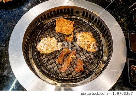 Various cuts of marinated meat and chicken grilling on a charcoal wire rack of a traditional Korean barbecue table. Overhead view of smoking savory food at a restaurant. Authentic Asian cuisine. 139708397