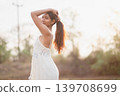 Side profile woman in white lace dress holding her long hair in warm sunset light, natural outdoor portrait for bohemian summer fashion, wellness, beauty, freedom, calm lifestyle, and travel. 139708699