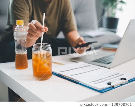 Iced tea glass and bottle on white desk with documents, smartphone, laptop, and blurred person, showing remote work refreshment, business break, productivity, and modern home office. Iced tea glass and bottle on white desk with documents, smartphone, laptop, and blurred person, showing remote work refreshment, business break, productivity, and modern home office. 139708761