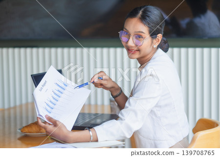 Smiling businesswoman in glasses points at a printed sales chart while presenting reports beside a laptop at a wooden meeting table, showing data analysis, teamwork, training, and success. Smiling businesswoman in glasses points at a printed sales chart while presenting reports beside a laptop at a wooden meeting table, showing data analysis, teamwork, training, and success. 139708765