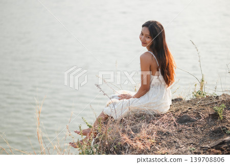 Smiling woman in white lace dress sitting beside calm lake and looking back in warm sunlight, showing relaxed summer vacation, peaceful nature, outdoor wellness, freedom, soft beauty, Smiling woman in white lace dress sitting beside calm lake and looking back in warm sunlight, showing relaxed summer vacation, peaceful nature, outdoor wellness, freedom, soft beauty, 139708806