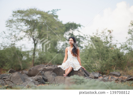 Woman in white lace dress sitting barefoot on dark rocks, surrounded by green trees and soft sky, peaceful outdoor portrait, bohemian summer fashion, nature wellness, travel, and calm relaxation. Woman in white lace dress sitting barefoot on dark rocks, surrounded by green trees and soft sky, peaceful outdoor portrait, bohemian summer fashion, nature wellness, travel, and calm relaxation. 139708812