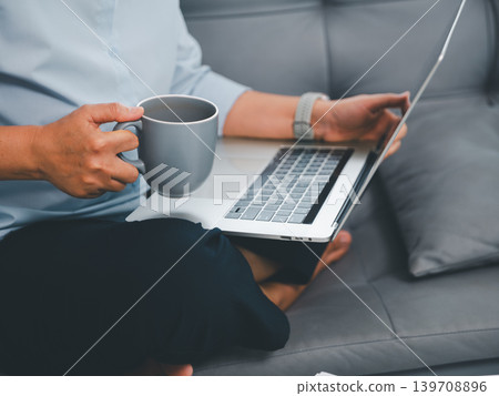 Close up professional holding gray coffee mug while using laptop on sofa, showing relaxed home office, remote work comfort, digital productivity, freelance business, and flexible lifestyle. Close up professional holding gray coffee mug while using laptop on sofa, showing relaxed home office, remote work comfort, digital productivity, freelance business, and flexible lifestyle. 139708896