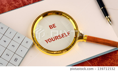 A golden magnifying glass highlights the words BE YOURSELF in red on white paper. A calculator and a dark fountain pen sit on a workspace, suggesting self-reflection A golden magnifying glass highlights the words BE YOURSELF in red on white paper. A calculator and a dark fountain pen sit on a workspace, suggesting self-reflection 139711532