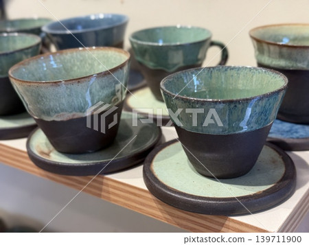 Ceramic cups with saucers displayed on shelf in pottery store, showcasing handcrafted design Ceramic cups with saucers displayed on shelf in pottery store, showcasing handcrafted design 139711900