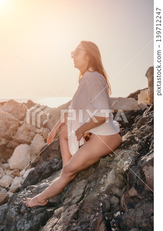 Woman beach sunset. Elegant mature woman in a white shirt sitting on rocky coastline during a golden hour sunset by the ocean. Woman beach sunset. Elegant mature woman in a white shirt sitting on rocky coastline during a golden hour sunset by the ocean. 139712247