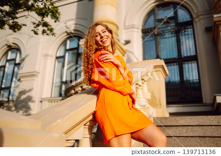 Young woman posing against backdrop of urban landscape in bright orange dress. Concept of lifestyle. 139713136