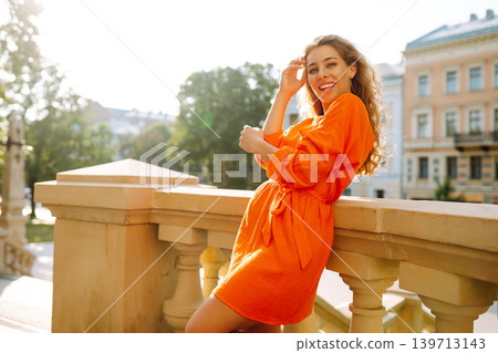 Young woman posing against backdrop of urban landscape in bright orange dress. Concept of lifestyle. 139713143