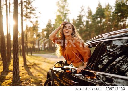 A young woman feels freedom leaning out of a car window in a sunny forest. Travel concept. A young woman feels freedom leaning out of a car window in a sunny forest. Travel concept. 139713430