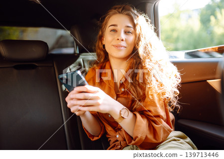 A young woman with a smartphone chats in the back seat of a car. Transport concept. Lifestyle. A young woman with a smartphone chats in the back seat of a car. Transport concept. Lifestyle. 139713446