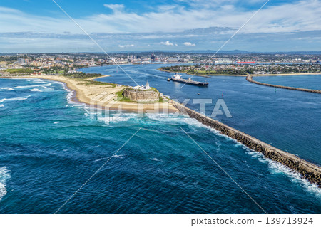 Aerial drone view of Nobbys Lighthouse and Nobbys Beach with a large cargo ship being escorted by tug boats through Newcastle Harbour in the background. Aerial drone view of Nobbys Lighthouse and Nobbys Beach with a large cargo ship being escorted by tug boats through Newcastle Harbour in the background. 139713924