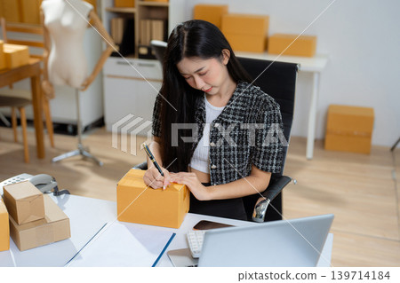 Asian vlogger woman influencer, SME retail store owner writing address on cardboard box at workplace. Asian vlogger woman influencer, SME retail store owner writing address on cardboard box at workplace. 139714184