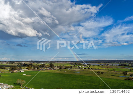 Aerial drone view of rural farmland in Maitland, New South Wales, Australia beneath cloudy blue sky with rainbow visible over agricultural landscape. Aerial drone view of rural farmland in Maitland, New South Wales, Australia beneath cloudy blue sky with rainbow visible over agricultural landscape. 139714492