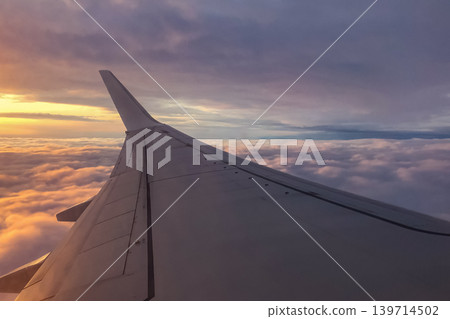 Airplane wing extends above thick clouds during evening flight. Soft sunset light reflects across the sky and aircraft surface. Airplane wing extends above thick clouds during evening flight. Soft sunset light reflects across the sky and aircraft surface. 139714502