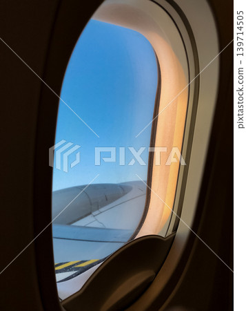 Airplane window frames clouds and wing during a calm evening flight. Soft sunset colors spread across the sky above the horizon. Airplane window frames clouds and wing during a calm evening flight. Soft sunset colors spread across the sky above the horizon. 139714505
