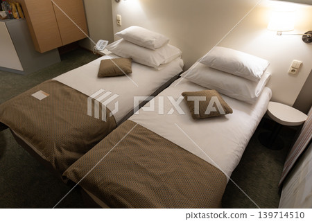 Twin beds with white linens and warm lighting fill a compact hotel bedroom. Neutral furniture and clean arrangement support comfortable overnight accommodation. 139714510