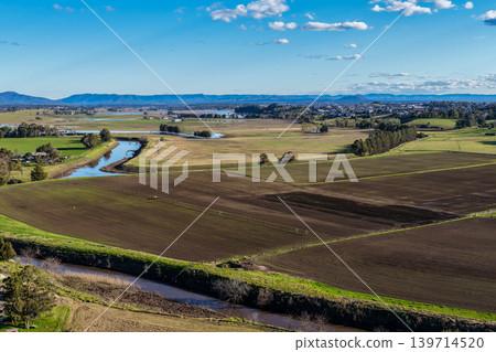 Aerial drone view of rural farmland in Maitland, New South Wales, Australia beneath cloudy blue sky with rainbow visible over agricultural landscape. Aerial drone view of rural farmland in Maitland, New South Wales, Australia beneath cloudy blue sky with rainbow visible over agricultural landscape. 139714520