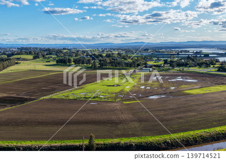 Aerial drone view of rural farmland in Maitland, New South Wales, Australia beneath cloudy blue sky with rainbow visible over agricultural landscape. Aerial drone view of rural farmland in Maitland, New South Wales, Australia beneath cloudy blue sky with rainbow visible over agricultural landscape. 139714521