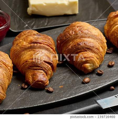 Golden croissants laid out in a row on a dark stone board, surrounded by coffee beans, butter, and raspberry jam Golden croissants laid out in a row on a dark stone board, surrounded by coffee beans, butter, and raspberry jam 139716460