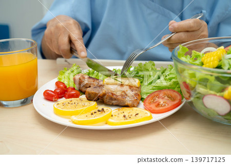 Asian elderly woman patient eating pork chop stake and vegetable salad for healthy food in hospital. Asian elderly woman patient eating pork chop stake and vegetable salad for healthy food in hospital. 139717125