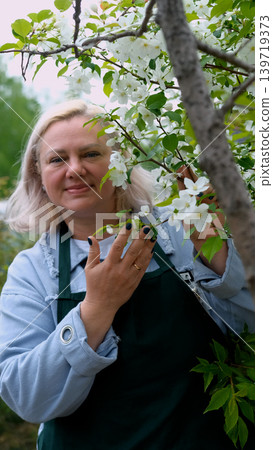 A portrait of a happy woman gardener, standing next to a blossoming apple tree, smiling, enjoying the pleasant scent of the flowers, and feeling happy. A portrait of a happy woman gardener, standing next to a blossoming apple tree, smiling, enjoying the pleasant scent of the flowers, and feeling happy. 139719373