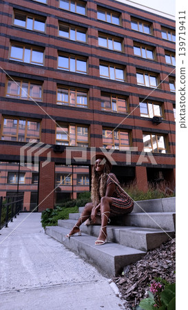 A young beautiful woman with afro-braids, wearing a brown hat, poses while sitting on the steps outside in an eco-friendly residential complex 139719411