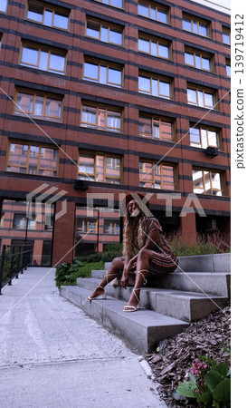 A young beautiful woman with afro-braids, wearing a brown hat, poses while sitting on the steps outside in an eco-friendly residential complex A young beautiful woman with afro-braids, wearing a brown hat, poses while sitting on the steps outside in an eco-friendly residential complex 139719412