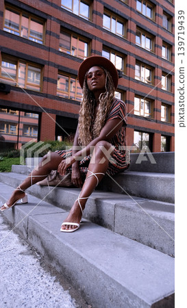 A young beautiful woman with afro-braids, wearing a brown hat, poses while sitting on the steps outside in an eco-friendly residential complex A young beautiful woman with afro-braids, wearing a brown hat, poses while sitting on the steps outside in an eco-friendly residential complex 139719439