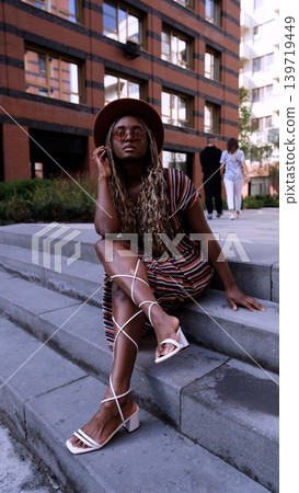 A young beautiful woman with afro-braids, wearing a brown hat, poses while sitting on the steps outside in an eco-friendly residential complex A young beautiful woman with afro-braids, wearing a brown hat, poses while sitting on the steps outside in an eco-friendly residential complex 139719449