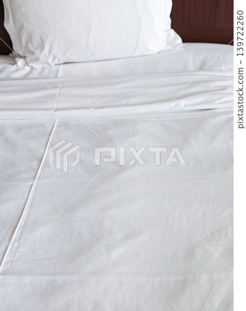 White bed linens with neatly arranged pillow on a bed in a hotel room, showcasing a clean and inviting sleeping environment White bed linens with neatly arranged pillow on a bed in a hotel room, showcasing a clean and inviting sleeping environment 139722260