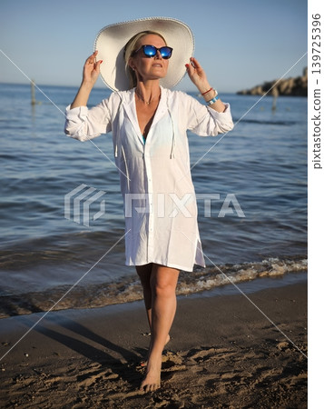 Woman in white tunic and wide-brimmed hat on sandy beach by the sea at sunset Woman in white tunic and wide-brimmed hat on sandy beach by the sea at sunset 139725396